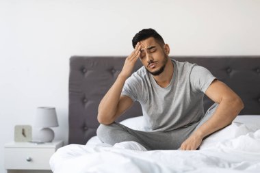 Genç bir Arap evdeki yatağının kenarında oturuyor, bitkin görünüyor. Acı dolu bir yüzü var. Yatak odasındaki yorgunluk ve rahatsızlıkla baş ederken başını tutuyor..