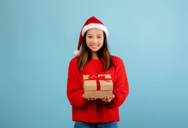 Genç bir kadın elinde paketlenmiş bir hediyeyle gülümsüyor. Kırmızı bir süveter ve Noel Baba şapkası giyer, parlak mavi bir arka plana bayram neşesi yayar..