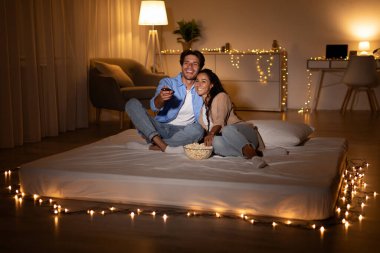 Sıcak bir oturma odasında, bir çift birlikte bir yatakta oturur, film gecesinin tadını çıkarırlar. Koca, elinde uzaktan kumandayla karısına sarılıyor. Yumuşak ışıklar rahat bir atmosfer yaratır..