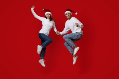 Mutlu çift el ele tutuşurken yükseğe zıplıyor, ikisi de Noel Baba şapkası ve günlük kıyafetler giyiyor. Parlak kırmızı arkaplan, o anın şenlikli ruhuna ekleniyor..