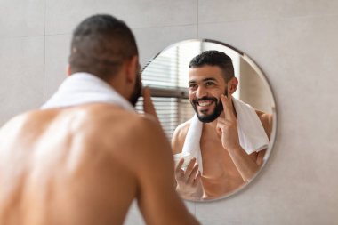 Genç Orta Doğulu adam aynada yüzüne nemlendirici sürerken gülümsüyor. Banyoda, boynunda beyaz bir havlu var. Neşeli tavırları kendine güveni gösteriyor..