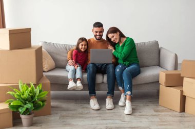 Gülümseyen Orta Doğulu aile, bir kanepede otururken dizüstü bilgisayarında oda tasarımı seçeneklerini araştırıyor. Etraflarını saran kutular ve bitkiler yeni yaşam alanlarını planlıyorlar..