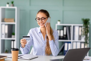 Mutlu bir iş kadını masasında oturmuş elinde kredi kartıyla akıllı telefonuyla konuşuyor. Ofis malzemeleri ile çevrili bir iş görüşmesi sırasında çevrimiçi ödeme sürecine katılmış..