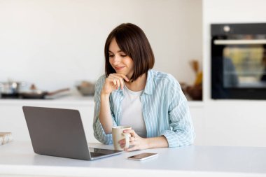 Bir kadın mutfak tezgahında oturuyor, elinde bir kahve fincanı var ve bilgisayarına odaklanmış. Parlak alan modern tasarım özelliklerini sergiliyor, üretkenlik için rahat bir atmosfer yaratıyor.