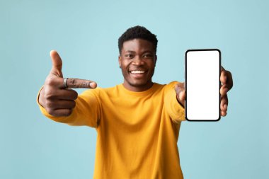 Sarı elbiseli yakışıklı Afro-Amerikan genç adam modern cep telefonunu işaret ediyor. Beyaz ekranı boş ve başparmağı mavi arka planda, model, en yeni mobil uygulamayı öneriyor.