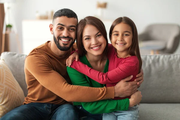 Üç kişilik neşeli bir Arap ailesi rahat kanepelerinde kucaklaşıyor. Sevgi dolu çift ve gülümseyen kızları bir anlığına mutlu bir şekilde evlerinde poz veriyorlar..