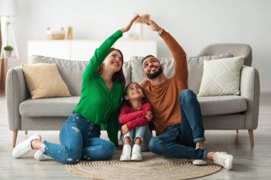 Sıcak bir oturma odasında, Arap ebeveynler sevinçle gülümseyen kızlarının üzerinde elleri olan bir çatı şekli oluştururlar. Birbirlerine yakın otururlar, kahkaha ve sevgiyi paylaşırlar..