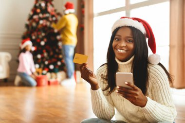 Sıcak kazaklı bir kadın yerde otururken telefonunu ve kartını tutuyor. Arka planda, aile üyeleri bir Noel ağacı süsler..