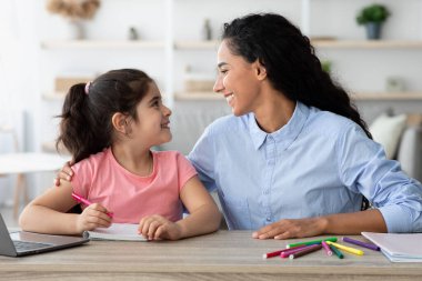 Bir anne ve küçük kızı ahşap bir masada birlikte çizerken neşeli bir anı paylaşıyorlar. Renkli işaretler etrafa saçılmış. Gülümseyip evde yaratıcı oyunlar oynuyorlar..