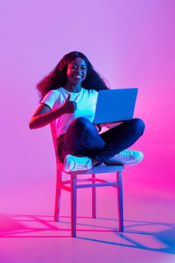 Neşeli genç siyah bir bayan bacak bacak bacak üstüne atmış bir sandalyede oturuyor ve dizüstü bilgisayarını kullanırken başparmağını kaldırıyor. Renkli neon ışıklar uzak araştırmaları teşvik eden enerjik bir atmosfer yaratır..