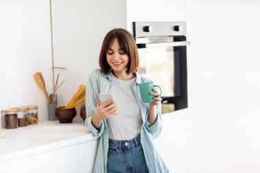 Genç bir kadın parlak bir mutfakta duruyor, bir elinde kahve fincanı, diğer elinde akıllı telefon tutarken gülümsüyor. Oda temiz, modern bir tasarıma sahip ve yemek aletleri görünür..