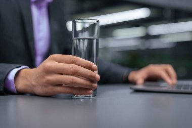 Bir işadamı ofisteki bir masada oturur. O bir elinde bir bardak su tutarken diğer elinde bir dizüstü bilgisayar kullanıyor. Çevre modern ve kapalı..
