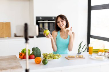 Neşeli genç bir kadın yeşil bir elma tutuyor ve parlak bir mutfakta onaylıyor. Taze meyve ve sebzeler tezgahta sergileniyor, sağlıklı bir yaşam tarzı sergileniyor..