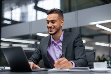 Takım elbiseli genç bir adam ofisteki bir masada oturuyor. O bir dizüstü bilgisayar üzerinde çalışıyor ve notlar alıyor. Doğal ışık boşluğu dolduruyor. Odaklanmış ve işine odaklanmış görünüyor..