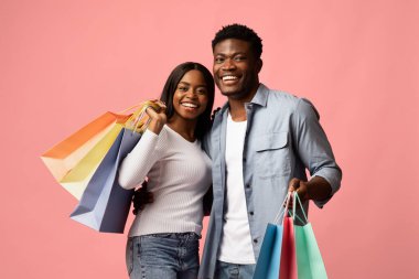 İki mutlu Afro-Amerikan aşığı ellerinde renkli alışveriş torbalarıyla gülümsüyor ve birbirlerine sarılıyorlar. Parlak pembe bir stüdyo geçmişleri var. Alışverişe olan sevgilerini sergiliyorlar..