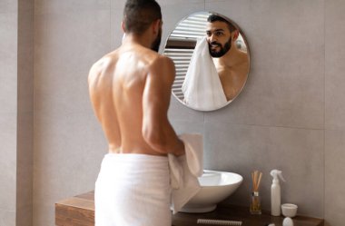Genç Arap adam modern bir banyoda duruyor, yuvarlak aynaya bakarken yüzünü silmek için havlu kullanıyor. Mutlu görünüyor ve günlük cilt bakımı ritüeliyle meşgul..