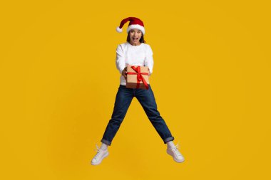 Noel Baba şapkası takan bir kız kırmızı kurdeleli bir hediye kutusunu tutarken sevinçle zıplıyor. Parlak sarı arka plan, bayram ruhunu kutlamak için şenlikli atmosfere ekleniyor..