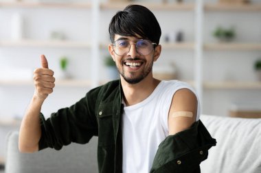 Genç bir adam evinde rahatça oturur, geniş bir şekilde gülümser ve tek koluyla baş parmağını kaldırır. Diğer kolunda yara bandı var. Aşısını orada yaptırmış..