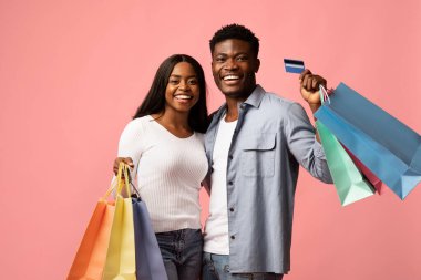 Genç siyah bir çift pembe bir arka plana karşı duruyor, alışveriş torbaları ve bir kredi kartı tutarken mutlu bir şekilde gülümsüyorlar. Birlikte başarılı bir alışveriş gezisini kutluyorlar..