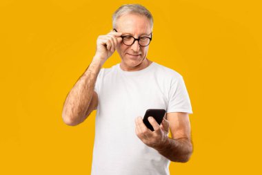 Bir adam parlak sarı bir arka planın önünde duruyor. O, elindeki akıllı telefonuna yakından bakarken gözlüğünü ayarlıyor. Ekrana odaklanmış görünüyor..