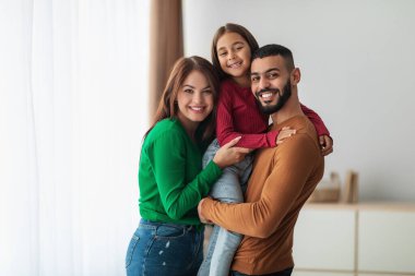 Sıcak bir oturma odasında, üç kişilik mutlu bir Arap ailesi keyifli bir anı paylaşıyor. Baba kızını kucaklarken anne de onları kucaklıyor ve kameraya sımsıkı gülümsüyor..