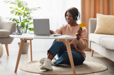 Bir kadın sıcak ve davetkar oturma odasında yerde otururken online alışveriş yapmaktan hoşlanır. Kulaklık takıyor ve elinde bir kart tutuyor. Dizüstü bilgisayarıyla meşgul..