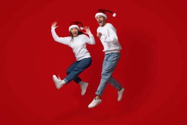 Noel Baba şapkalı iki neşeli kişi atlayıp canlı kırmızı bir arkaplana karşı kutlama yapıyorlar. Noel sezonu boyunca neşe ve tatil ruhunu ifade ediyorlar..