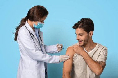 Doktor önlüğü uzmanı sakin bir ortamda genç bir adama aşı veriyor. Her ikisi de maske takıyor, güvenli sağlık uygulamaları sergiliyor. Ortam dostane ve profesyonel görünüyor..