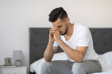 Üzgün, düşünceli, orta doğulu bir adam evinde yatağının kenarında oturuyor, başını ellerinin üzerinde dinlendiriyor. Düşünceli ve üzgün görünüyor, hayat zorluklarını ya da mevsimsel depresyonu yansıtıyor..