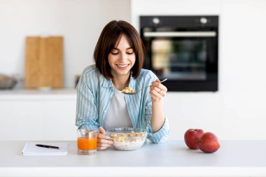 Genç bir kadın parlak bir mutfakta mısır gevreği yerken gülümsüyor. Yakınlarda bir bardak portakal suyu ve taze elmalar var. Neşeli bir sabah ortamı yaratıyor..