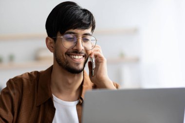 Gözlüklü genç bir adam telefonda konuşurken gülümsüyor. Dizüstü bilgisayarına çok az dekorasyonla aydınlık bir ofis alanına odaklanmış..