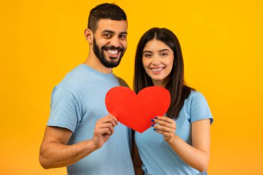Mutlu bir Arap çift, kameraya gülümseyen kırmızı bir kalbe sarılıyor. Sevgililer Günü 'nde birbirlerine uyumlu mavi tişörtler giyip, parlak sarı bir arkaplanda aşklarını sergiliyorlar..