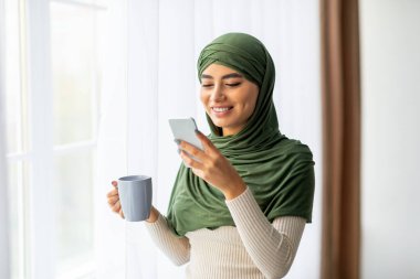 Tesettürlü genç bir Arap kadın elinde sıcak bir fincan kahveyle pencerenin yanında duruyor. Akıllı telefonuna göz atarken, sohbet ederken ve çevrimiçi iletişim kurarken gülümsüyor. Sabah ışığı sıcacık boşluğu doldurur.