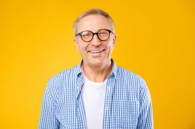 Kocaman bir gülümsemeyle kameraya bakan bir adam. Gözlük takıyor ve beyaz bir gömleğin üstüne işaretli bir gömlek giyiyor. Arka plan parlak sarı. Neşeli ve rahat görünüyor..