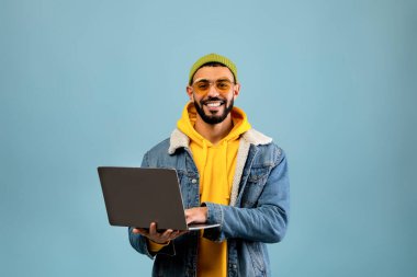 Sarı kapüşonlu ve kot ceketli neşeli genç bir adam dizüstü bilgisayar kullanıyor. Şık bir güneş gözlüğü ve yeşil bir bere takıyor. Arkaplan koyu mavi renkte, canlı bir sahne yaratıyor..
