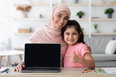 Neşeli bir anne ve kızı bir dizüstü bilgisayarla tahta bir masada oturuyorlar. Bitkiler ve okul malzemeleriyle dolu iyi ışıklandırılmış bir odadalar. Birlikte vakit geçirmenin keyfini çıkarıyorlar..