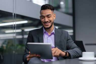 Ofisteki bir masada oturan bir adam gülümseyerek tablete bakıyor. Takım elbise giyiyor ve işiyle meşgul görünüyor. Ofisin büyük pencereleri ve modern bir tasarımı var..