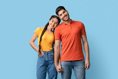 İki genç yetişkin el ele tutuşurken mutlu mesut gülümsüyor. Parlak renkli gömlekler giyiyorlar, mavi arka planda neşeli bir atmosfer yaratıyorlar..