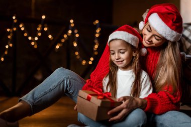 Bir anne ve küçük kızı rahat bir ortamda yerde oturuyorlar. Kırmızı tatil kazakları ve Noel Baba şapkaları giyiyorlar..