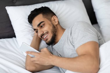Yakışıklı bir Arap 'ın yatakta rahat bir şekilde dinlenirken, video izlemekten ve akıllı telefonuyla mesajlaşmaktan zevk alırken, evde modern teknolojiyi kucaklarken yakın plan fotoğrafı..