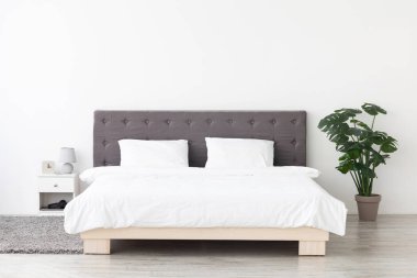 Bu geniş yatak odasında beyaz çarşaf giymiş büyük, rahat bir çift kişilik yatak sergileniyor. Minimalist dekor, modern mobilyalar ve tencerede yeşil bir bitki içererek rahat bir atmosfer yaratıyor..