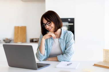 Kahverengi saçlı ve gözlüklü bir kadın mutfak masasında oturup dizüstü bilgisayarına odaklanıyor. Mutlu ve meşgul görünüyor. Etrafı temiz ve parlak mutfak düzeniyle çevrili..