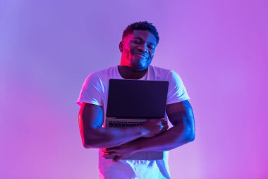 Neşeli genç siyah adam canlı neon ışıklarda dururken boş bir ekranla dizüstü bilgisayara sarılıyor. Bu sahne teknolojiyle ilgili reklamlar veya web sitesi tasarımları için harika bir zemin sunar..