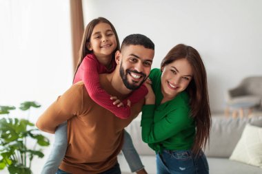 Neşeli ebeveynler parlak oturma odalarında kızlarıyla güzel vakit geçiriyorlar. Her iki ebeveyn de mutlu bir şekilde gülümserken çocuk mutlu bir şekilde babasının sırtında dönüyor..