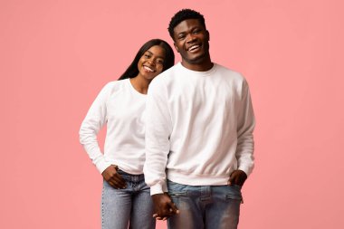 Genç Afrikalı Amerikalı çift pembe bir stüdyoda yan yana duruyor. Elele tutuşup gülümsüyorlar, kadın başını erkeğin omzuna yaslıyor, neşeli bir an yaratıyor..