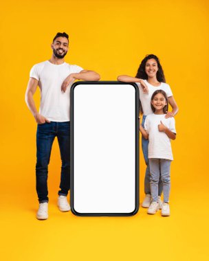 Üç kişilik bir aile, geniş ve boş bir akıllı telefonun her iki tarafında aileleri olan canlı sarı bir arkaplan karşısında duruyor. Sevinç saçan çocuk başparmağını kaldırır..