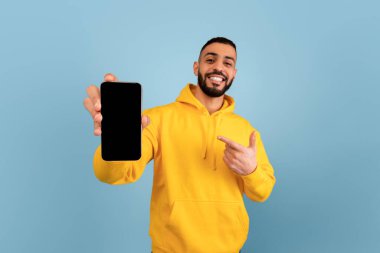 Gülümseyen genç bir adam parlak sarı bir kapüşon giyerken akıllı telefonunu işaret ediyor. Arka plan pürüzsüz mavi, neşeli bir atmosfer yaratıyor..