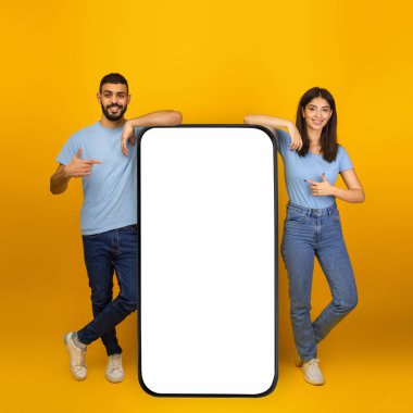 Genç bir Arap çift, boş ekranlı büyük bir akıllı telefonu hevesle işaret ederek birlikte duruyorlar..