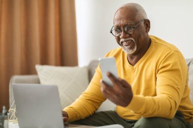 Yaşlı bir adam gözlük takıyor ve akıllı telefonunu kontrol ederken gülümsüyor. Yakındaki bir dizüstü bilgisayarla rahat bir şekilde bir kanepede oturuyor. İyi aydınlatılmış bir yaşam alanında. Teknolojiyle eğleniyor..