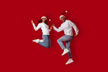 Neşeli bir çift heyecanla zıplıyor, Noel Baba şapkaları ve günlük kıyafetler giyiyorlar. Canlı kırmızı bir arka planda tatil ruhlarını gösteriyorlar..